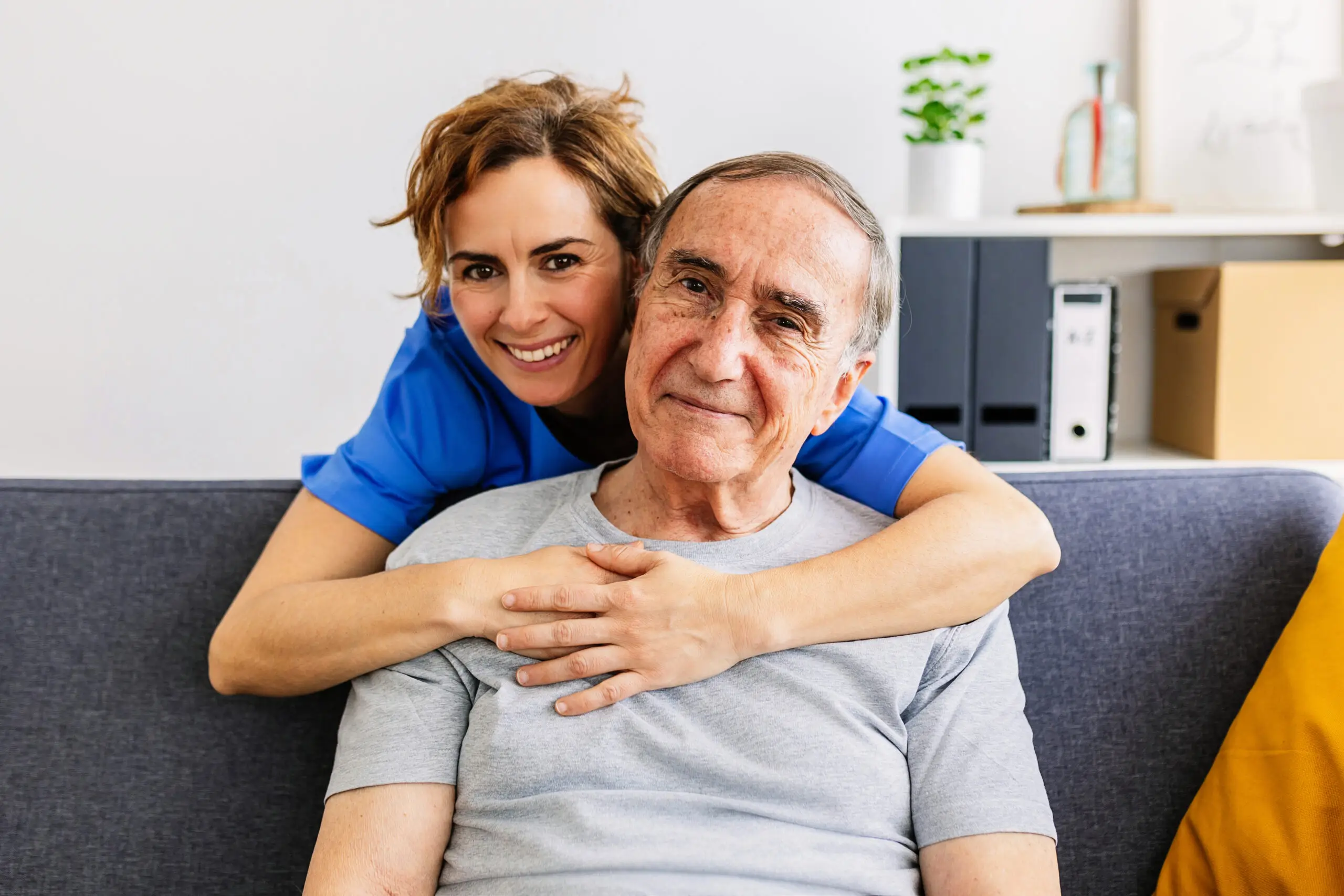 A caregiver in blue uniform smiles and embraces an older man sitting on a sofa at home.