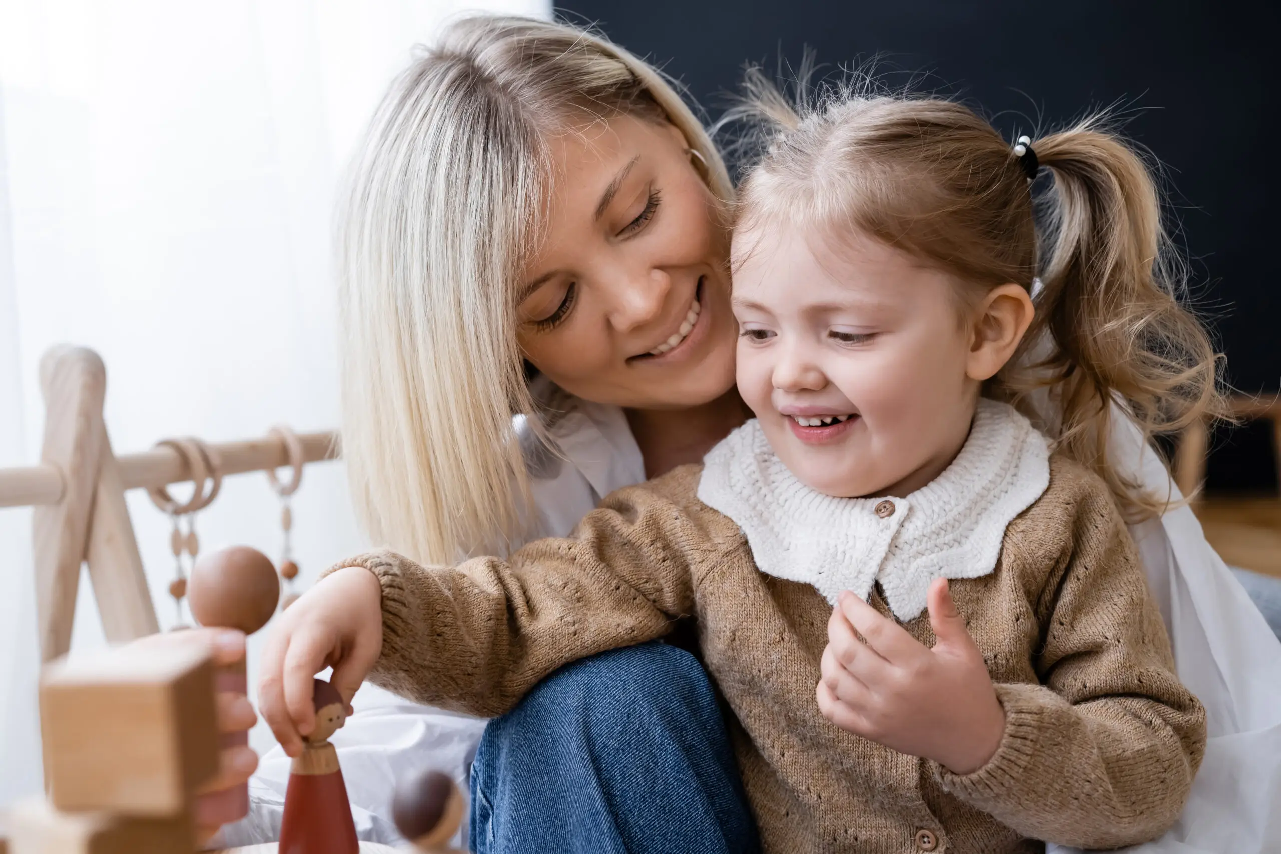 A woman and a young girl smile while playing together with wooden toys at home.