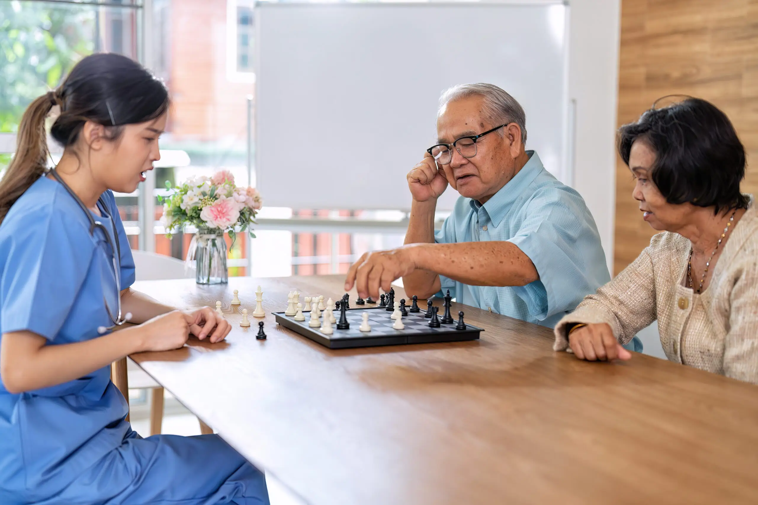 A care worker in scrubs plays chess at a table with an older couple.