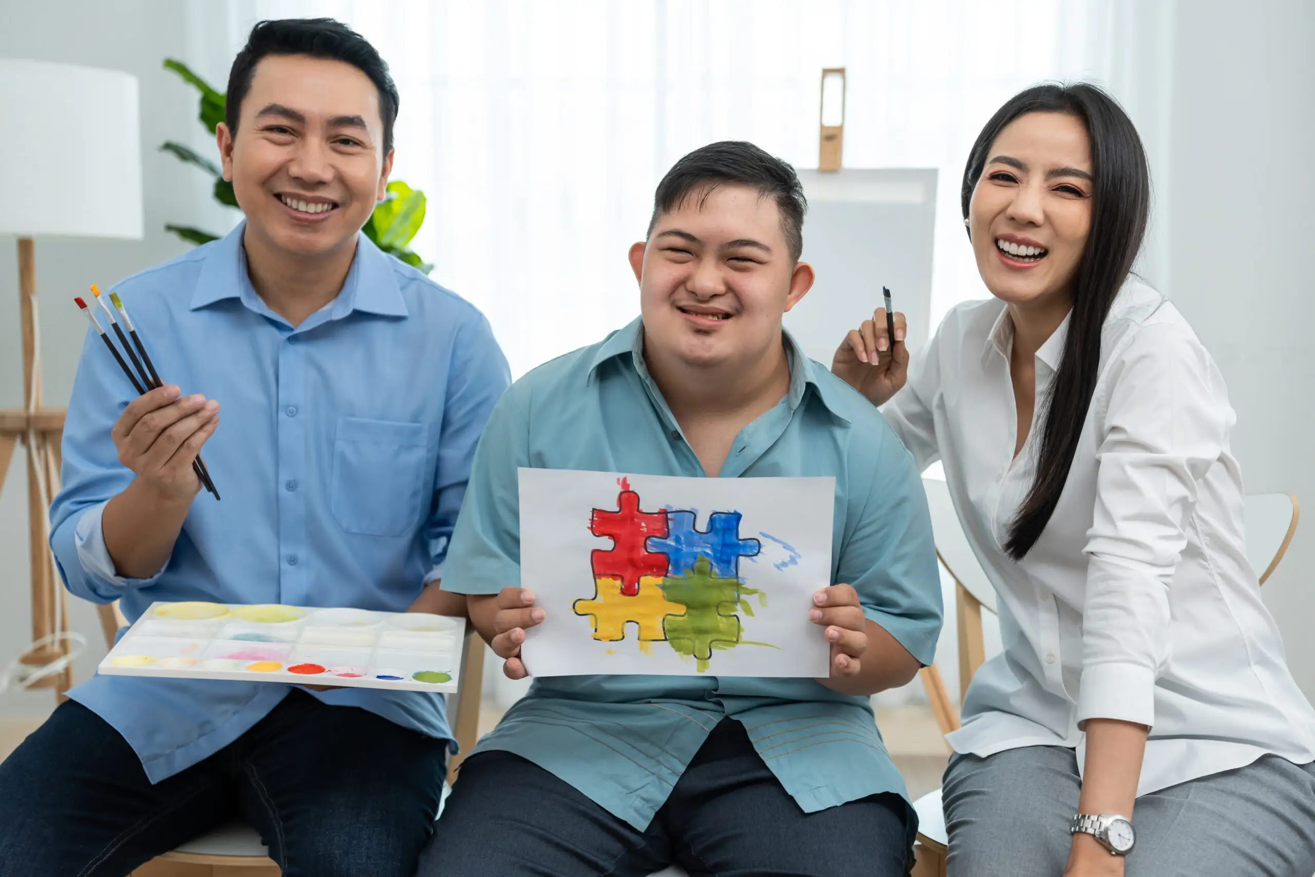 A young person holds a colourful puzzle painting while sitting between two smiling adults with art supplies.