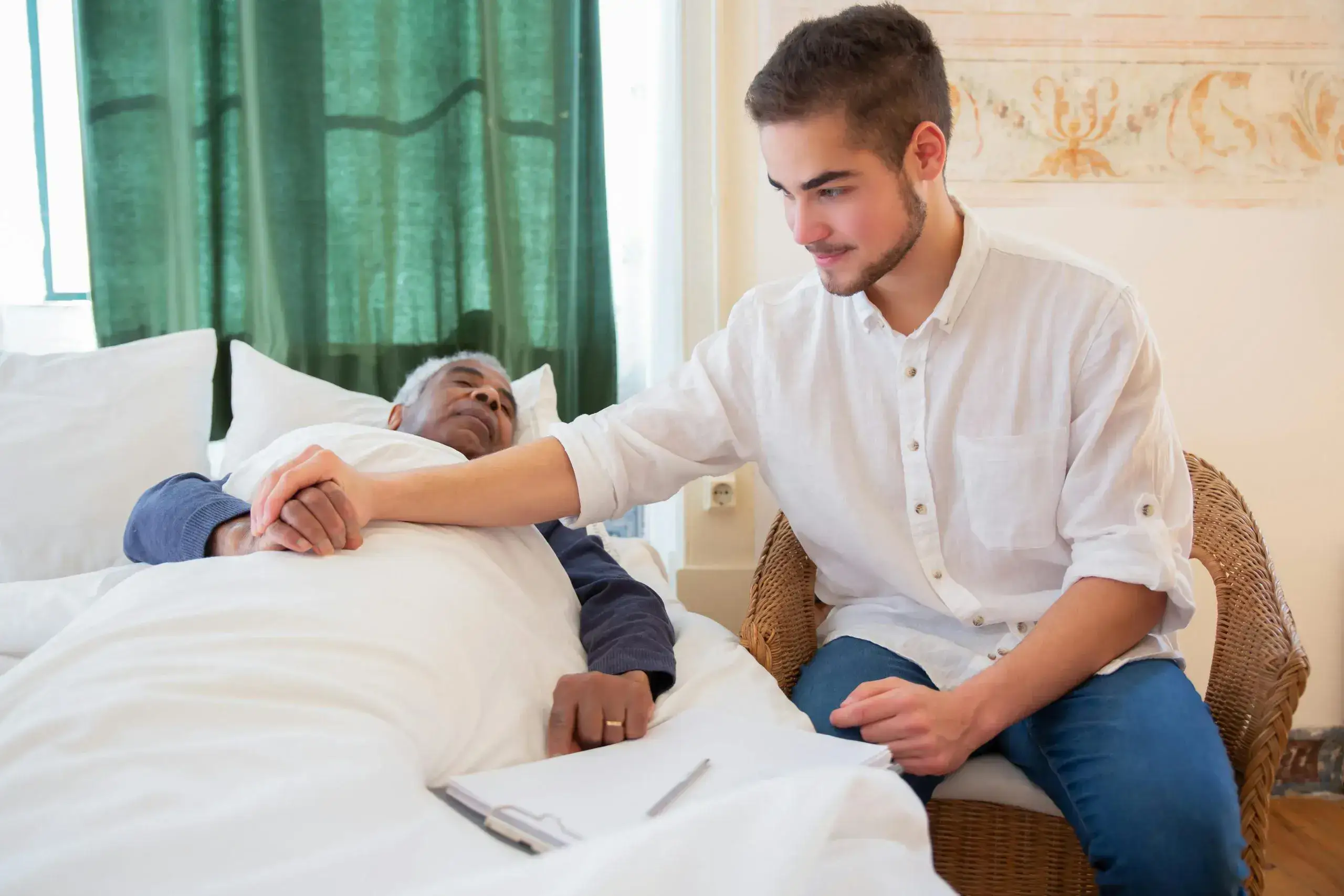 A support worker sits beside an older man resting in bed and holds his hand during a home visit.
