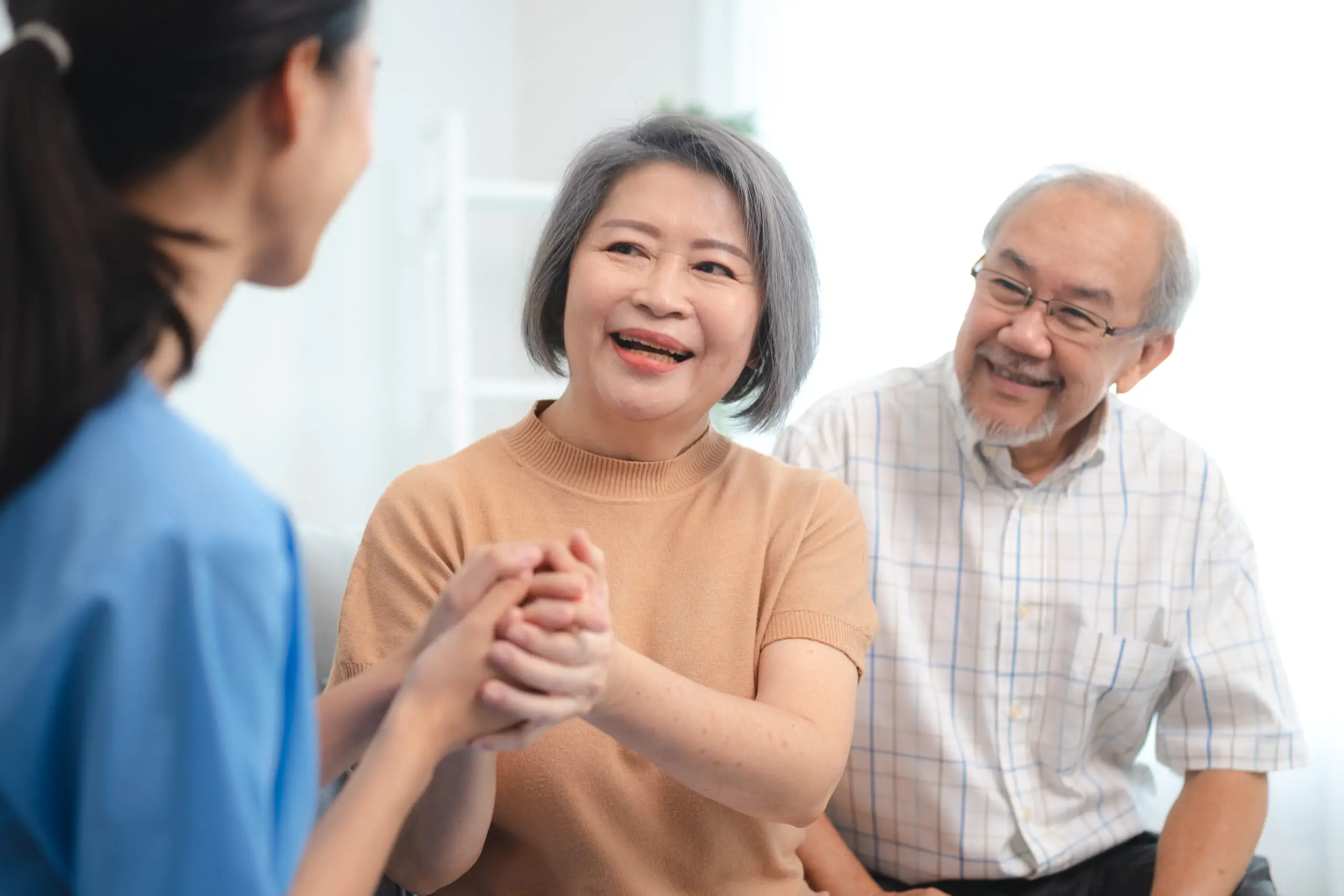 A care worker holds the hands of a smiling older woman while an older man sits beside them during a home visit.