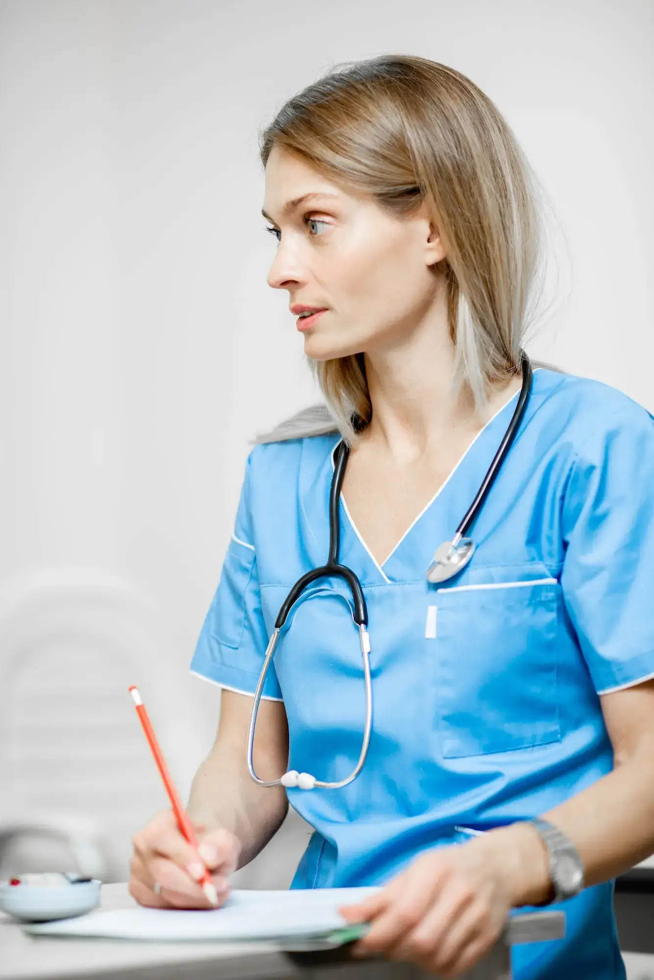 A care professional in blue scrubs writes notes while holding a clipboard.