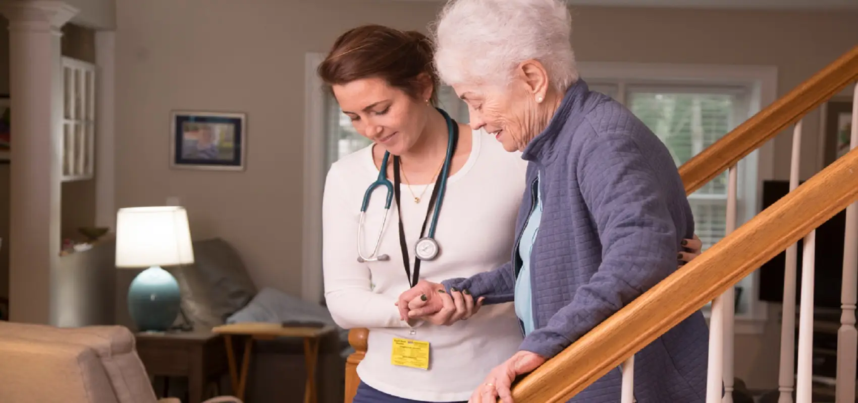 A care worker steadies an older woman by the arm as they walk down the stairs at home.